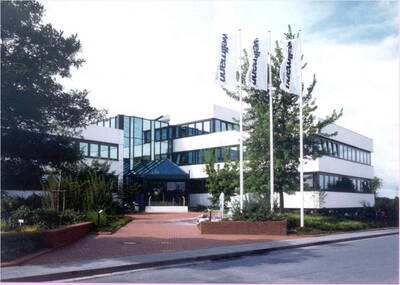 Modernes Bürogebäude mit großflächigen Glasfronten, bepflanztem Eingangsbereich und drei weißen Flaggenmasten an der Straße
