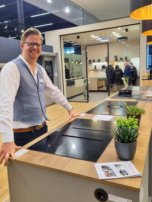 Lächelnder Aussteller präsentiert an einem Messestand eine moderne Kücheninsel mit integriertem Kochfeld und Dunstabzug, während im Hintergrund Besucher eine offene Musterküche besichtigen.
