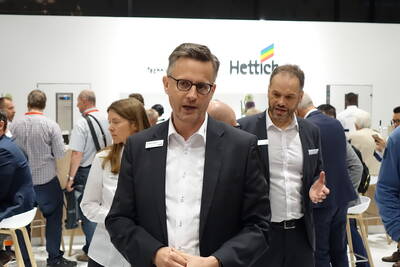 Geschäftsmann im Anzug spricht auf einer gut besuchten Messe vor dem Hettich‑Stand mit weiteren Fachbesuchern im Hintergrund