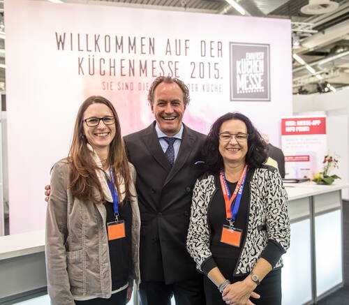 Drei lächelnde Messemitarbeitende mit Namensschildern posieren gemeinsam vor dem Willkommensbanner der Küchenmesse 2015 am Informationsstand.