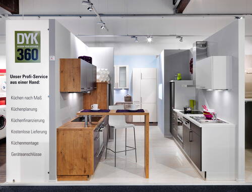 Modernes Küchenstudio mit zwei Musterküchen, Holzelementen, weiß-grauen Fronten, Kochfeld mit Töpfen und Servicewand mit Leistungen wie Küchenplanung und Montage
