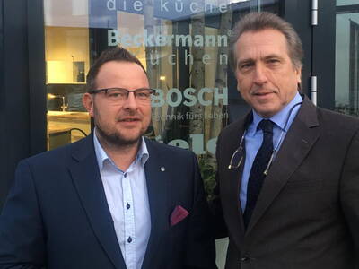 Zwei Männer in Anzügen stehen vor einer Glasfassade mit Küchen- und Bosch-Logo und blicken in die Kamera