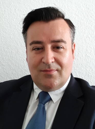 Mann im dunklen Anzug mit blau gestreifter Krawatte blickt ruhig in die Kamera vor einer hellen, strukturierten Wand.