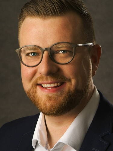 Lächelnder Mann mit Brille, kurzem Bart und Anzug vor dunklem Hintergrund in einem professionellen Porträt.