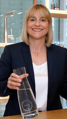 Lächelnde Frau im Business-Anzug hält eine Glaskaraffe mit Wasser in einer modernen Küchen- oder Ausstellungsumgebung