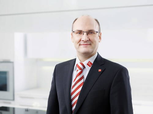 Geschäftsmann mit Brille im dunklen Anzug und rot-weiß gestreifter Krawatte lächelt vor einem hellen, modernen Bürohintergrund.