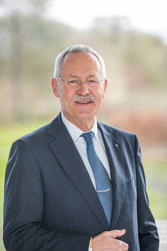 Älterer Mann im dunklen Anzug mit blauer Krawatte und Brille steht lächelnd vor einem unscharfen, grünen Hintergrund im Freien.