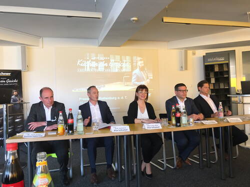 Fünf Geschäftsleute sitzen bei einer Pressekonferenz nebeneinander an einem Tisch mit Namensschildern und Getränkeflaschen, während im Hintergrund eine Präsentation zur Marke „Lechner“ projiziert wird.