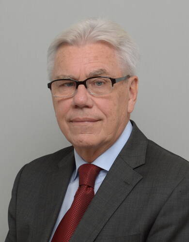 Älterer Mann mit weißem Haar, Brille und dunklem Anzug mit roter Krawatte blickt ruhig in die Kamera vor neutralgrauem Hintergrund.