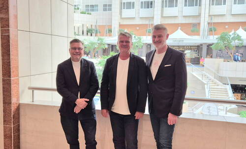 Drei lächelnde Männer in dunklen Blazern und weißen T‑Shirts stehen nebeneinander in einer hellen, modernen Atrium‑Einkaufspassage mit Treppen, Café und Pflanzen im Hintergrund.