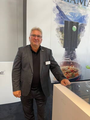 Mittelalterlicher Mann im grauen Anzug steht lächelnd an einem Messestand vor einem großformatigen Produktplakat mit Berglandschaft im Hintergrund