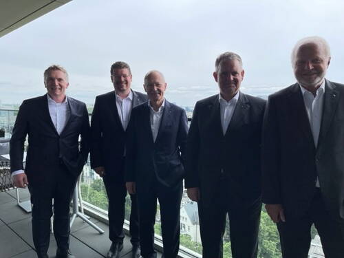 Fünf Männer in dunklen Anzügen stehen lächelnd auf einer Terrasse mit Glasbrüstung, im Hintergrund eine Stadtlandschaft unter bewölktem Himmel.