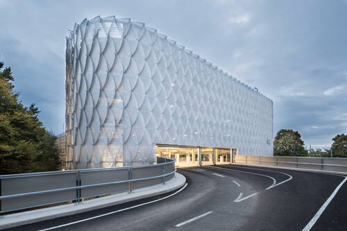 Geschwungen ansteigende Zufahrtsrampe führt zu einem modernen Parkhaus mit lichtdurchlässiger, schuppenartiger Fassade aus überlappenden weißen Elementen bei bewölktem Himmel