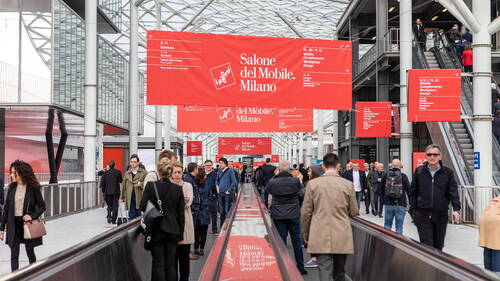 Besucherinnen und Besucher gehen über ein überdachtes Messegelände mit roten Bannern des Salone del Mobile Milano, während Rollsteige und Rolltreppen sie durch die moderne Glas- und Stahlhalle führen.