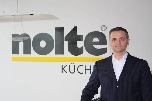 Geschäftsmann im dunklen Nadelstreifenanzug steht vor einer weißen Wand mit großem „nolte Küchen“-Logo.