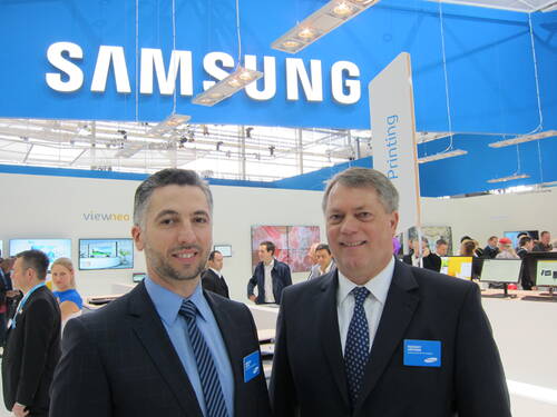 Zwei Geschäftsleute in Anzügen stehen lächelnd vor einem belebten Samsung-Messestand mit Monitoren und Besuchern im Hintergrund.