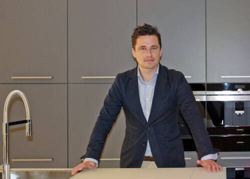 Mann im Sakko lehnt sich in moderner grauer Einbauküche an die Arbeitsplatte und blickt direkt in die Kamera