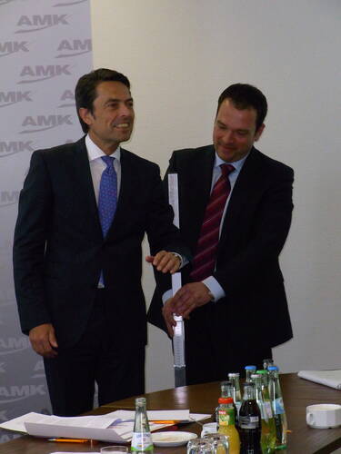 Zwei Geschäftsleute im Anzug lächeln und halten gemeinsam ein schmales, längliches Objekt in einem Konferenzraum, vor ihnen ein Tisch mit Getränkeflaschen, Gläsern und Unterlagen.