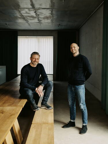 Zwei lässig gekleidete Männer in Jeans und dunklen Pullovern posieren entspannt in einem modernen, minimalistisch eingerichteten Raum mit langem Holztisch und Betonwänden.
