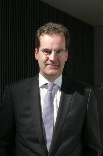 Mittelalter Mann im dunklen Anzug mit helllila Krawatte und Brille steht vor einer dunklen, geriffelten Wand und blickt ruhig in die Kamera