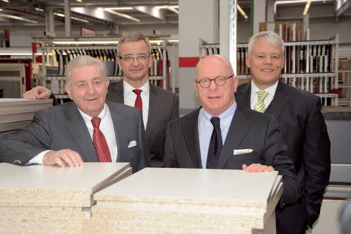 Vier Geschäftsleute im Anzug stehen in einer Produktionshalle hinter gestapelten Holz- und Spanplatten und blicken in die Kamera