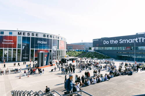 Menschenmengen versammeln sich vor dem Eingang der Messe Berlin zur IFA, umgeben von großen Werbebannern und modernen Messegebäuden bei sonnigem Wetter.