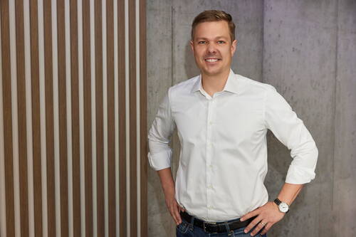 Mann in weißem Hemd steht lächelnd mit Händen in der Hüfte vor einer Wand aus Holzlamellen und Beton.