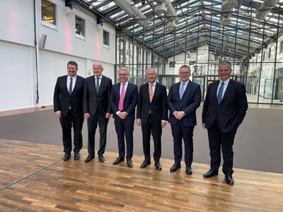 Sechs elegant gekleidete Männer in Anzügen posieren lächelnd in einer modernen, lichtdurchfluteten Industriehalle mit Glasdach und Holzfußboden.