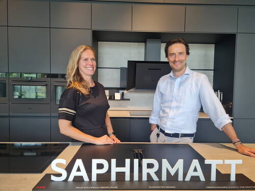 Zwei lächelnde Personen präsentieren in einer modernen, dunkel gehaltenen Einbauküche ein Kochfeld mit der leuchtenden Aufschrift „SAPHIRMATT“.