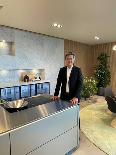 Eleganter Geschäftsmann steht an einer modernen Edelstahl-Kochinsel in einem stilvoll eingerichteten Showroom mit Marmorrückwand und großen Zimmerpflanzen