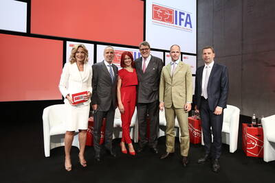 Sechs elegant gekleidete Fachleute posieren auf einer IFA‑Bühne vor großen Bildschirmen mit dem Messe‑Logo und weißen Sesseln im Hintergrund