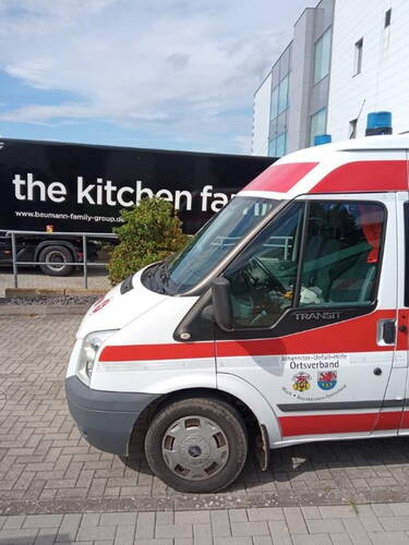 Rettungswagen der Johanniter-Unfall-Hilfe parkt auf einem gepflasterten Parkplatz vor einem schwarzen Lkw mit der Aufschrift „the kitchen family“ und einem modernen Bürogebäude im Hintergrund.