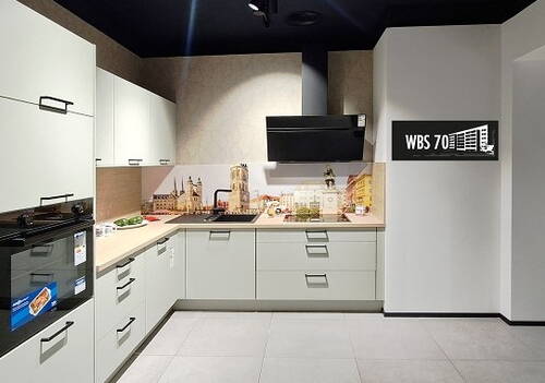 Moderne Einbauküche in L‑Form mit hellen Fronten, Wandfoto einer europäischen Altstadt über der Arbeitsplatte und schwarzer Dunstabzugshaube vor dunkler Decke