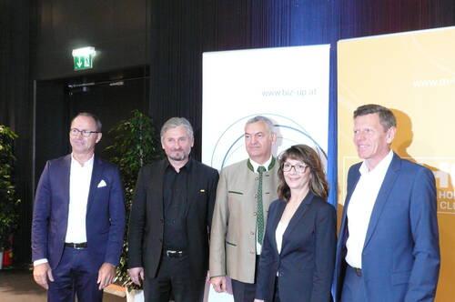 Fünf Geschäftsleute posieren in formeller Kleidung vor Messe- und Firmenbannern in einem hell beleuchteten Konferenzraum.