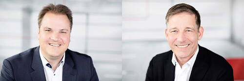 Zwei lächelnde Geschäftsmänner im Anzug nebeneinander in einem hellen, modernen Büro porträtiert