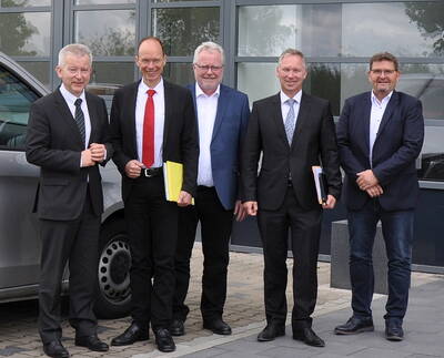 Fünf Männer in Anzügen stehen lächelnd nebeneinander vor einem Firmengebäude und halten Unterlagen in der Hand
